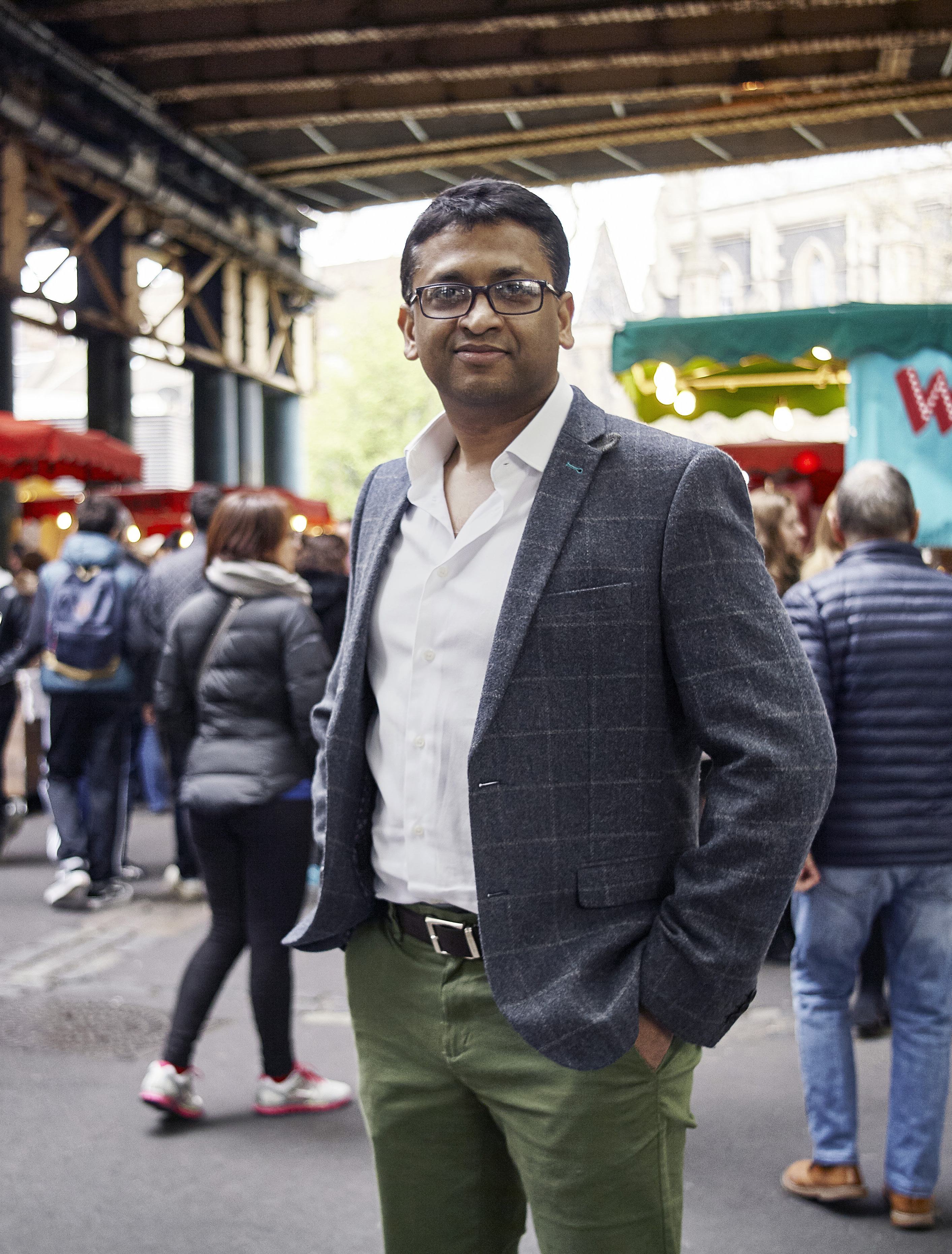 Portrait of a man in glasses and a gray jacket, standing in an outdoor market with people and stalls in the background.
