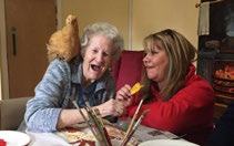 Elderly woman laughing with a chicken on her shoulder, next to a smiling younger woman at a table with art supplies.