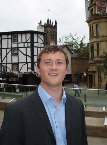 Portrait of a man in a dark suit and light blue shirt, standing outdoors in a city with historic buildings.