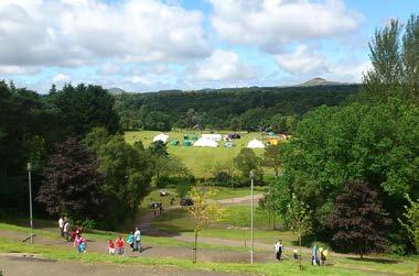 An aerial view of an outdoor field with white tents, surrounded by trees and hills. People walk on a path in the foreground.