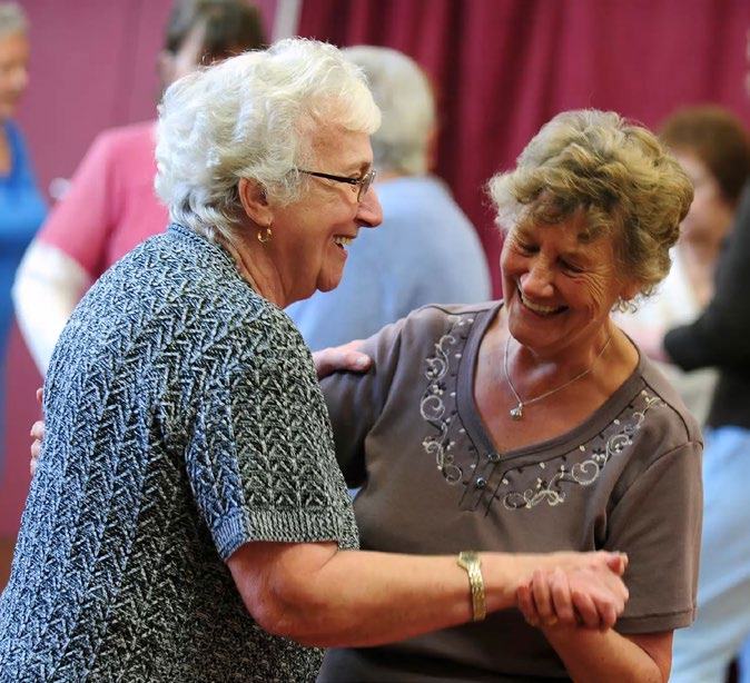 Two elderly women laugh and dance together joyfully in a social setting, with other people in the background.