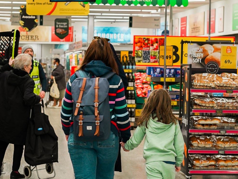 Shoppers, including a woman and child, in a busy supermarket aisle with shelves of baked goods, produce, and various price signs.