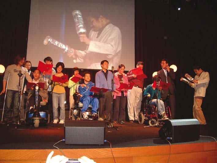 A group of people, some in wheelchairs, on a stage performing, with a large screen behind them showing a man holding a bottle.