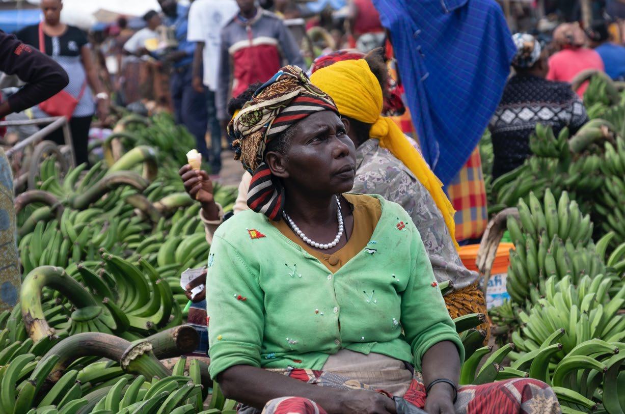 Woman in a market surrounded by green bananas, wearing a patterned headwrap and pearl necklace, with other vendors in the background.
