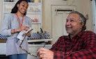 A young woman with a clipboard talking to an older man smiling at a table in a home setting.