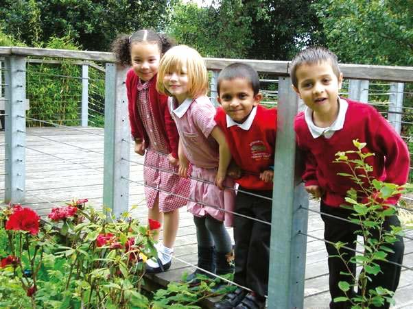 Four diverse young children in school uniforms smiling while peeking over a railing outdoors among red flowers.
