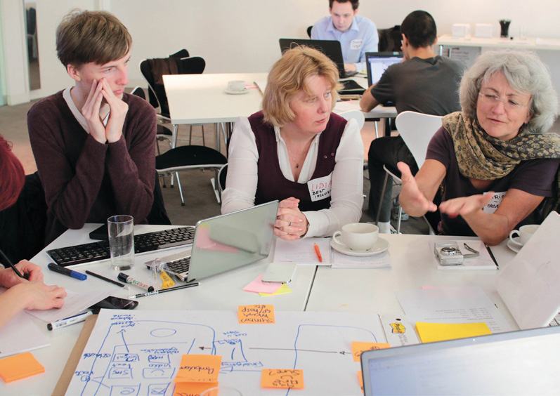 People collaborating at a table with laptops, a whiteboard diagram, and sticky notes illustrating a conceptual model or process.