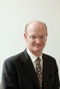 Headshot of a smiling bald man wearing glasses, a dark suit, and a polka-dotted tie, looking directly at the camera.