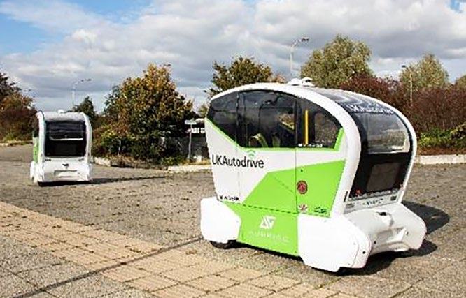 Two white and green "UK Autodrive" self-driving shuttle buses on a paved lot under a partly cloudy sky.
