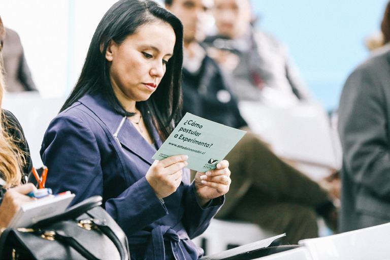 Woman reading a flyer asking "¿Cómo postular a Experimenta?" (How to apply to Experimenta?) at an event.