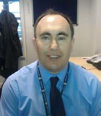 Headshot of a man in a blue shirt, tie, and lanyard in an office.