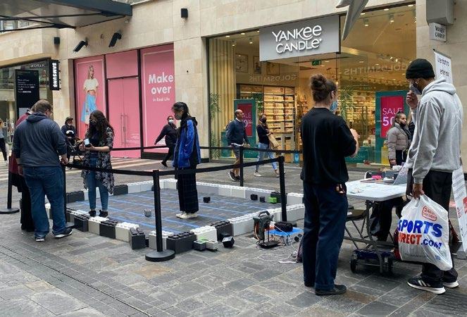 People gathered around an interactive display made of large blocks in a shopping mall with store fronts in the background.