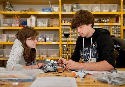 A young man and woman working on assembling a small robot or mechanical device on a table, with shelves of components in the background.