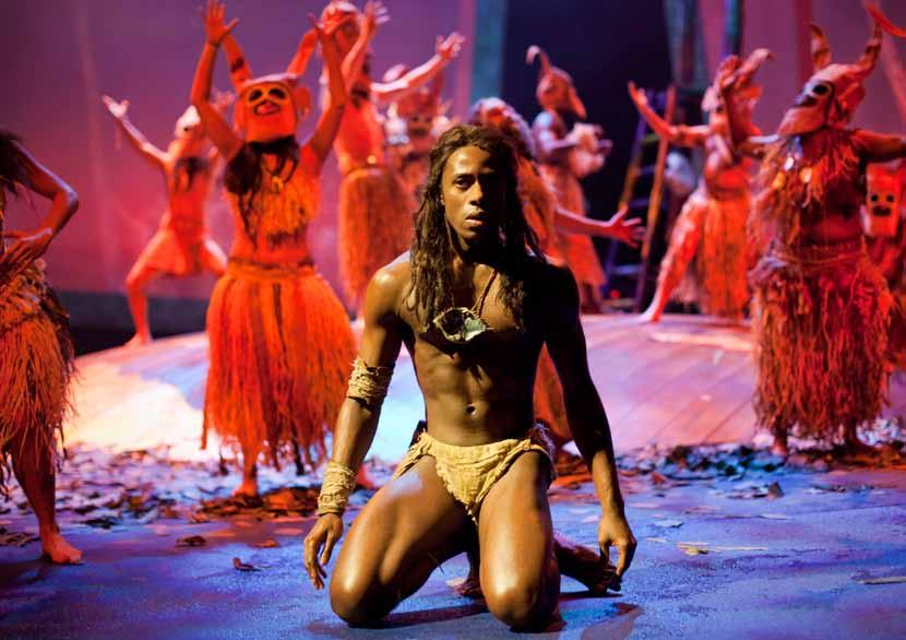 A man with long dark hair and minimal costume kneels center stage, looking forward. Other costumed performers are blurred in the background.