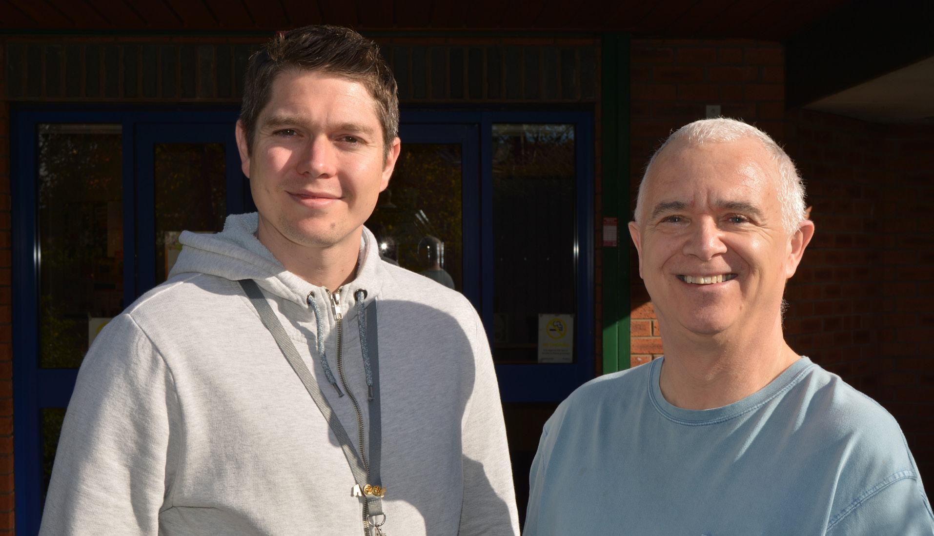 Two men smiling, standing outdoors in front of a brick building.