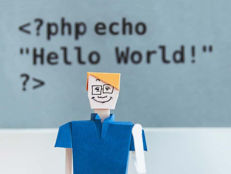 An origami figure stands in front of a blurred background displaying the PHP code <?php echo "Hello World!"; ?>, representing programming or development.