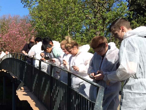 Group of people in white hazmat suits standing on a metal bridge, looking at their phones or devices.