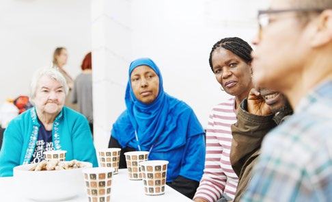 Diverse group of people, mostly women, sitting around a table with cups and snacks, engaged in conversation.
