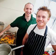 Two smiling men in a kitchen, one wearing a striped apron, with cooking pots and a tray of roasted vegetables visible in the foreground.