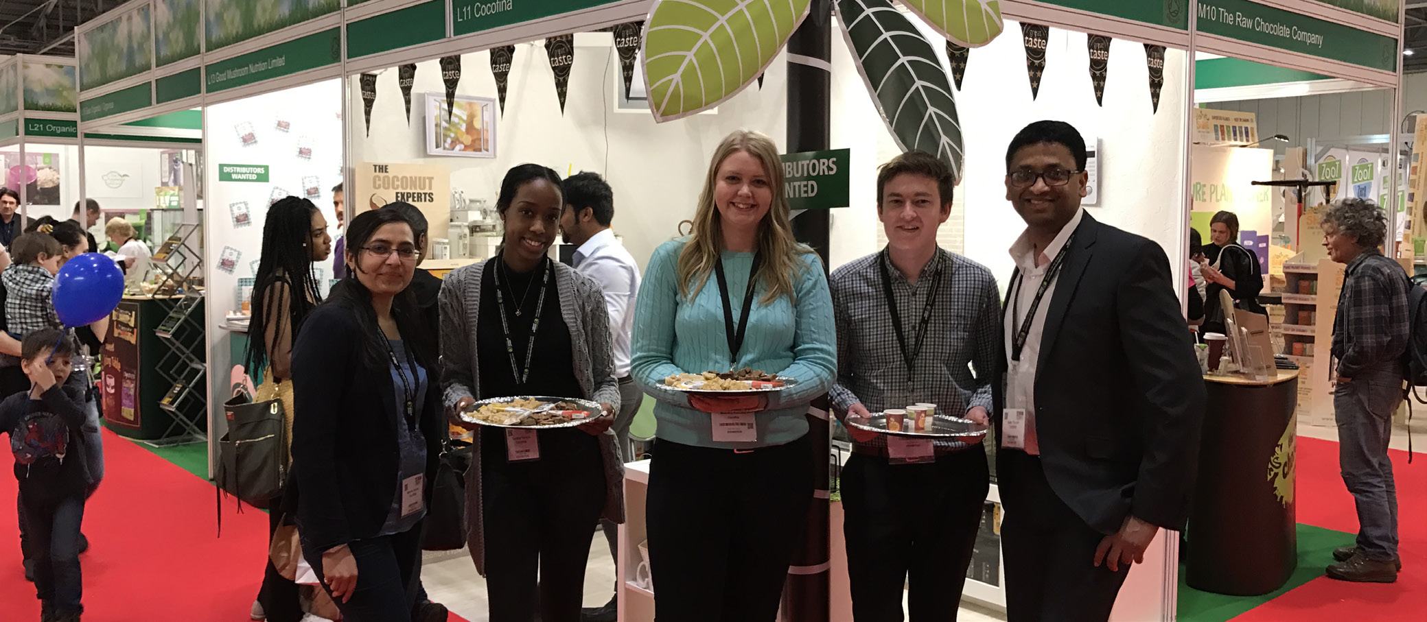 Five people, four holding food sample trays, stand in front of "The Coconut Experts" booth at a trade show or exhibition.