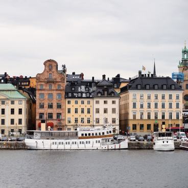 Waterfront view of historic buildings in a city, with two boats docked along the quay under a cloudy sky.