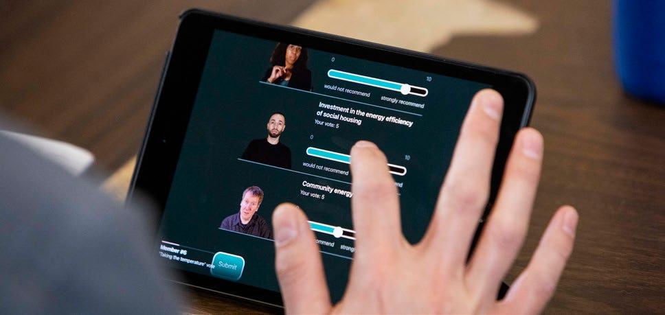 A person's hand interacts with a tablet displaying an online survey with sliders for voting on topics like investment in social housing and community energy.