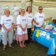 Four adults wearing white t-shirts standing behind a blue-covered table with various promotional materials at an outdoor event under a white tent.