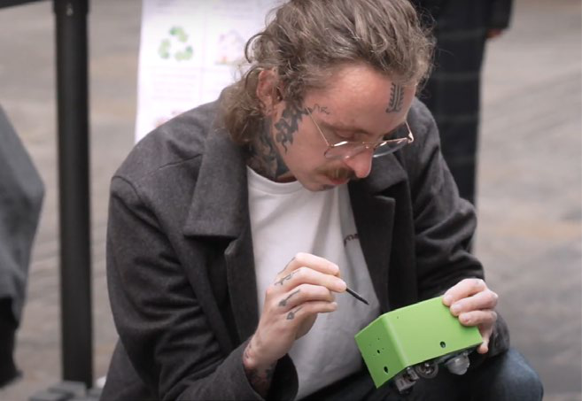 Close-up of a person with tattoos and glasses, focused on assembling a small green electronic device.