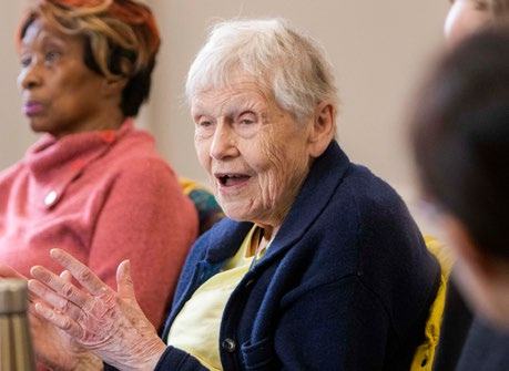 An elderly woman with white hair and a blue cardigan speaks animatedly in a group setting, gesturing with her hands.