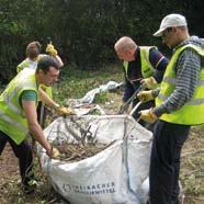 A group of people in high-visibility vests and gloves filling a large white sack with branches and debris during an outdoor clean-up activity.