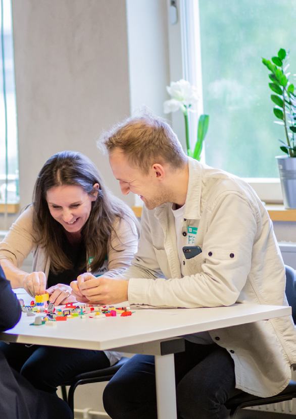 Two people laughing while building with colorful LEGO bricks at a table, depicting a creative workshop.