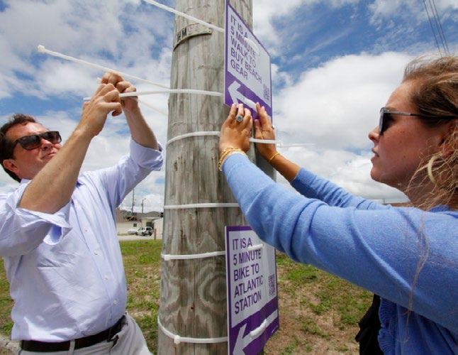 Two people attaching purple informational signs with QR codes to a wooden utility pole using white zip ties.