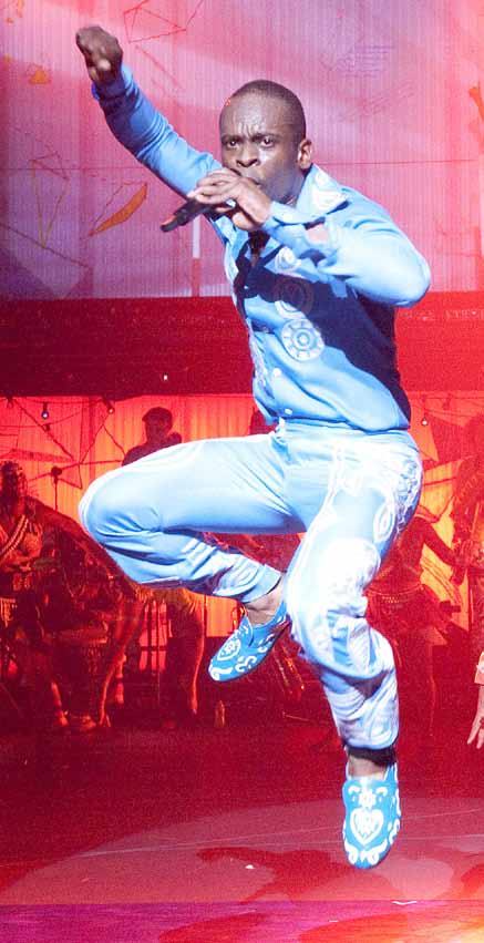 A male dancer in a light blue patterned suit leaps mid-air on a stage with red lighting.