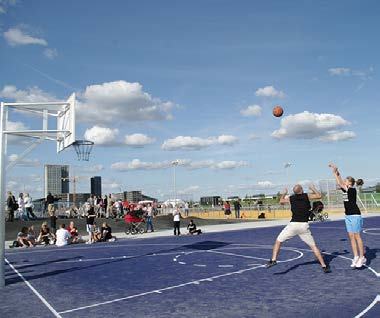Two people playing basketball on an outdoor blue court with onlookers in the background under a partly cloudy sky.