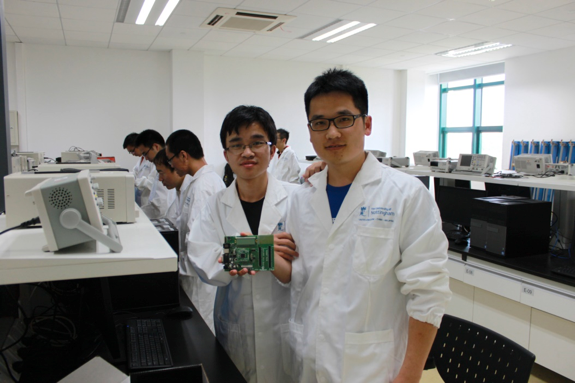 Two students in lab coats holding a circuit board in a lab, with other lab equipment and students visible.