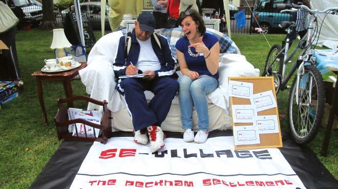 Man and woman at an outdoor "SE-VILLAGE THE PECKHAM SETTLEMENT" stall with a corkboard, bike, and sofa.