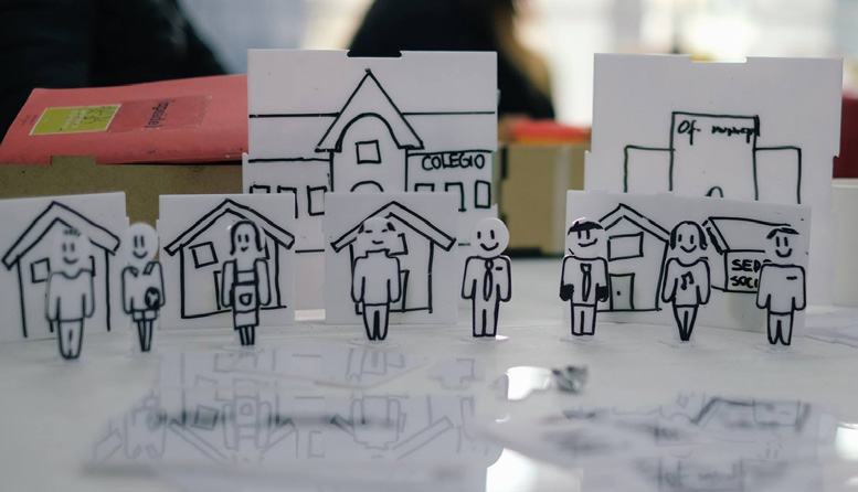 Paper cutouts illustrating stick figures representing people and simple line drawings of buildings, including houses, a school, and an office.