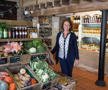 A woman smiling in a farm shop with fresh vegetables and other products displayed around her.