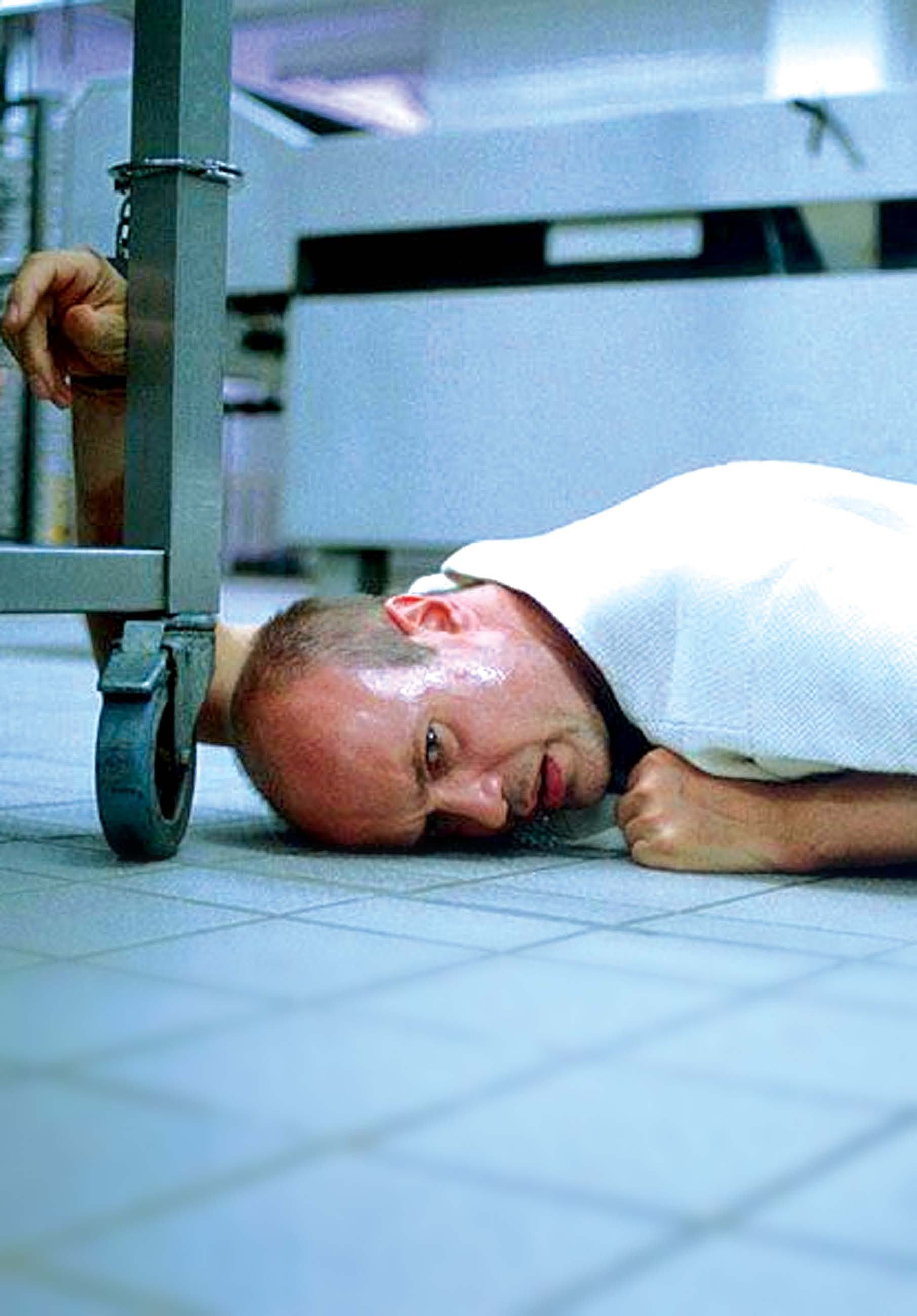 A man with a bald head lies on a tiled floor, his head near a metal cart wheel, looking up distressed.