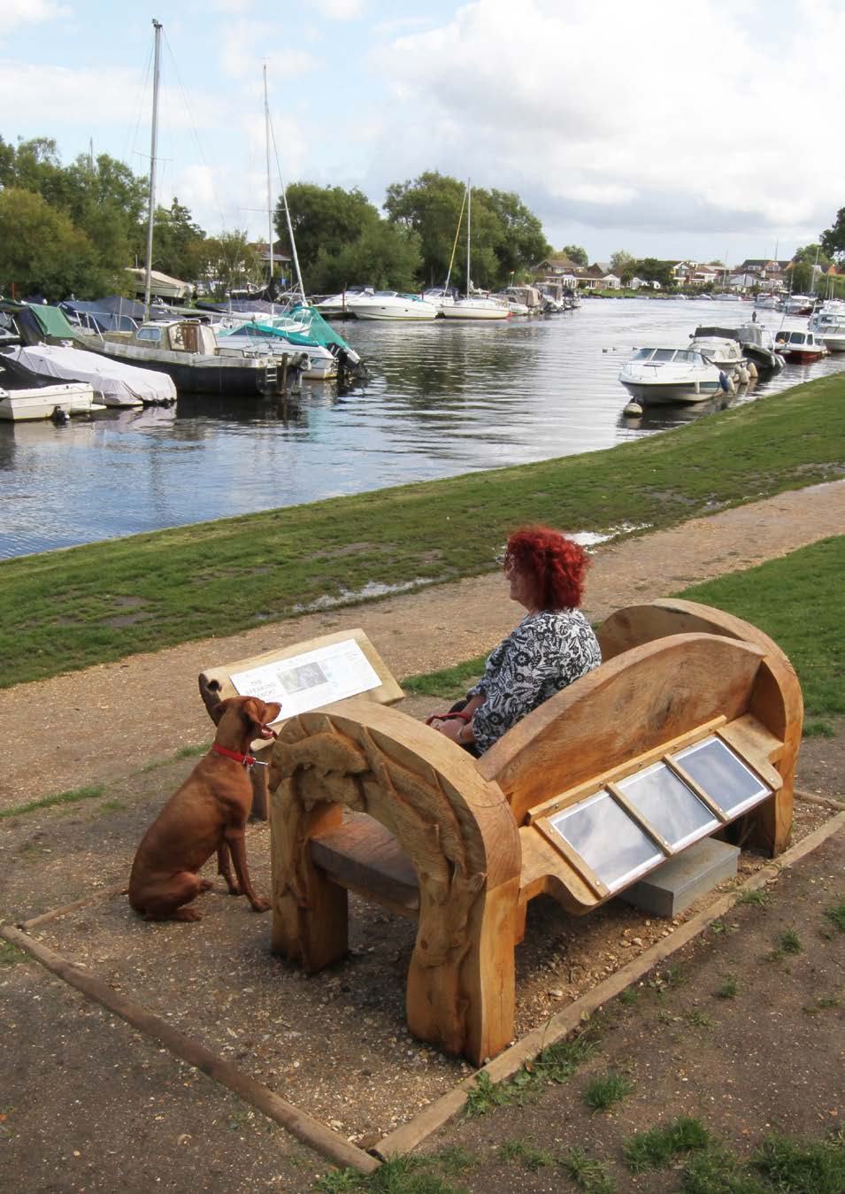 Woman with a dog sitting on a carved wooden bench overlooking a river with boats, next to an informational sign.