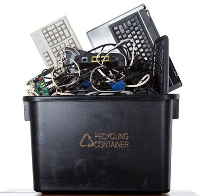 A black recycling container overflowing with old electronic waste, including keyboards, laptops, and cables.
