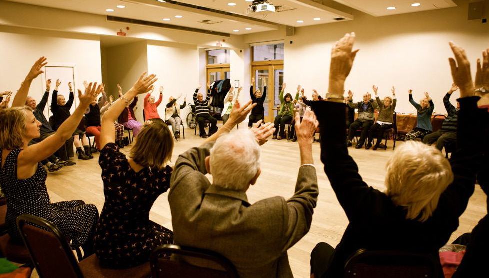 A group of diverse people, including many seniors, sitting with their arms raised during an activity in a brightly lit room.