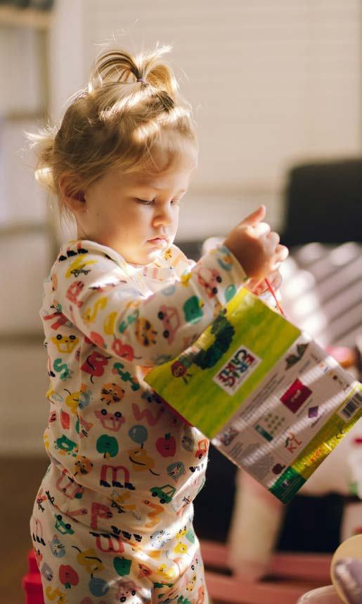 A toddler in patterned pajamas holds and looks at a small green picture book in bright sunlight.