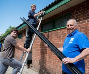 Three men installing a new gutter system on a brick building, with one on a ladder holding tools and two others holding a long gutter section.