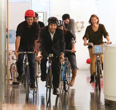 Employees joyfully ride bicycles through an office hallway, possibly part of a workplace wellness initiative or event.