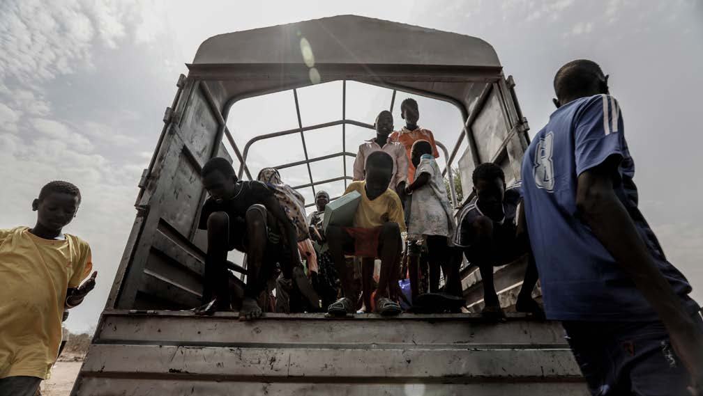 Group of children and adults standing crowded in the back of an open-top truck under an overcast sky.