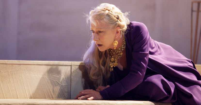 An actress in a purple dress and gold jewelry, likely Helen Mirren, leans forward on a stage looking distressed.