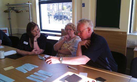 Three people, two women and a man, engaged in a discussion at a table with cards and papers, in a meeting setting.
