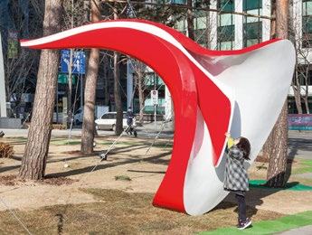 A child stands next to a large, undulating red and white abstract sculpture in a park, with trees and buildings behind.