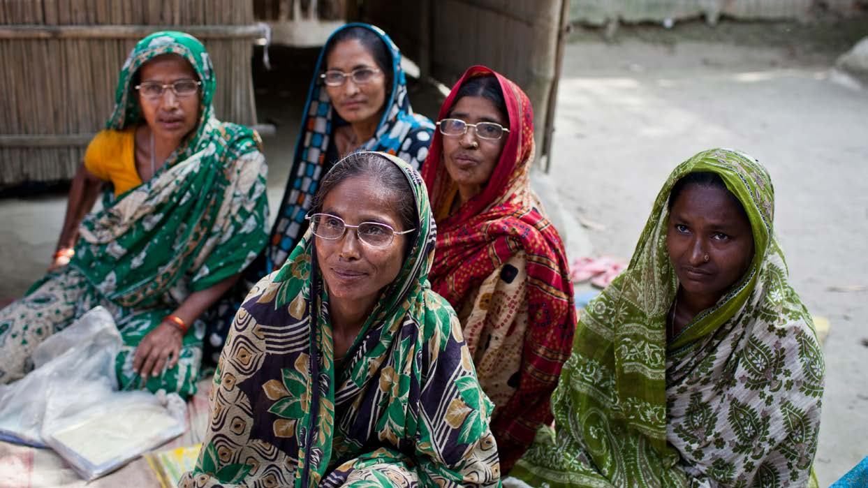 Five women in traditional South Asian clothing sit together outdoors, possibly representing beneficiaries of a community program.